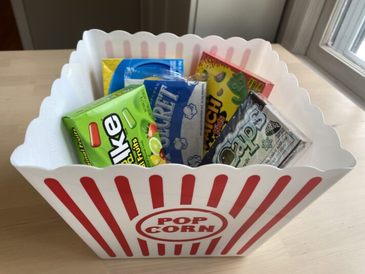 A picture showing a red and white classic striped popcorn tub and various boxes of movie candy and a bag of microwavable popcorn inside