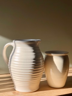 Two locally made vases. Both are cream colored. The one in the foreground has a horizontal texture and a handle while the one in the background is smooth with a small rim.